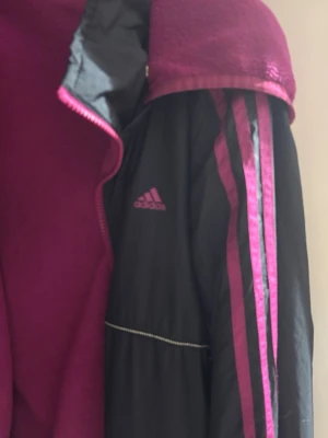 Svart och cerise Adidas vindjacka - Snygg svart vindjacka från Adidas med cerise detaljer och fleecefoder. Jackan har klassiska Adidas-ränder på ärmarna, dragkedja framtill och huva med mjukt fleecefoder. Perfekt för blåsiga dagar och sportiga outfits.