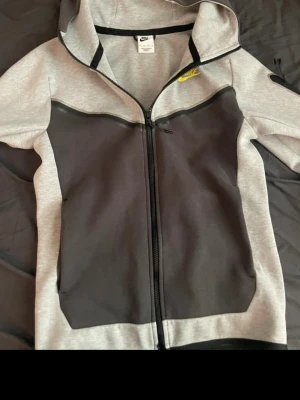 Nike grå och svart hoodie XS - Nike hoodie i grått och svart med dragkedja framtill och gul Nike-logga på bröstet. Hoodien har huva och är tillverkad i mjukt material, perfekt för en sportig och avslappnad stil. Snygg kontrast mellan färgerna och klassisk passform.
