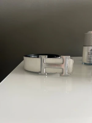 Vändbart svart och vitt skärp med H-spänne - Snyggt vändbart bälte i svart och vitt med ett stort H-format spänne i silver. Bältet är gjort i skinn och har en stilren design som passar till många olika outfits. Perfekt accessoar för att lyfta din look.