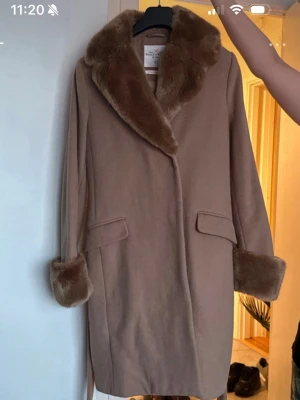 Beige kappa med fuskpälsdetaljer - Stilren beige kappa med bred krage och muddar i mjuk fuskpäls. Kappan har två fickor framtill och ett bälte i midjan för snygg passform. Perfekt för kyliga dagar när du vill vara både varm och trendig.