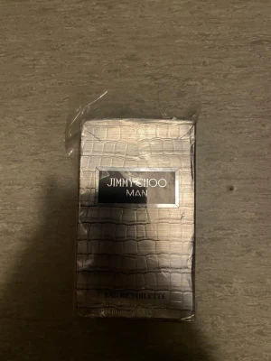 Jimmy Choo Man Eau de Toilette - Säljer en Jimmy Choo Man Eau de Toilette. Förpackningen har ett stilrent silverfärgat krokodilskinnsmönster och svart etikett med vit text. Parfymen är för män och kommer i en klassisk rektangulär kartong.