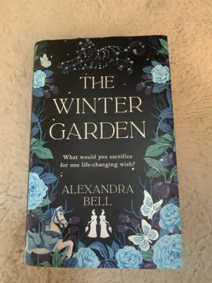 The Winter Garden av Alexandra Bell - En magisk och fantasifull roman med hård pärm, svart omslag dekorerat med blå rosor, fjärilar och en stjärnbeströdd panter. Boken handlar om en ovanlig tävling i en förtrollad trädgård där priset är en livsförändrande önskan. Perfekt för dig som gillar fantasy och äventyr.