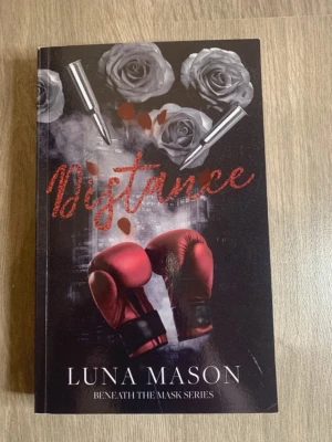 Distance av Luna Mason - Snygg pocketbok med svart omslag, röda boxningshandskar, grå rosor och patroner på framsidan. Titeln 'Distance' står i röd glittrig text och författaren är Luna Mason. Tillhör serien 'Beneath the Mask Series'. Perfekt för dig som gillar dramatik och spänning.