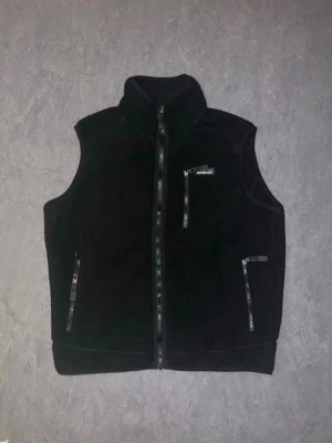 Svart Patagonia Väst - Snygg svart dunväst med hög krage och hel dragkedja framtill. Västen har tre fickor med dragkedja, varav en på bröstet. Tillverkad i ett mjukt, fluffigt material som ger en sportig look. Perfekt att slänga över en hoodie eller tröja för extra värme.