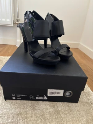 United nude heels - Snygga svarta pumps med hög klack och bred rem över foten. Skorna har öppen tå och en modern, edgy design med blanka detaljer. Perfekta för dig som vill sticka ut och addera en cool touch till din outfit.