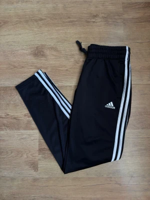 Svarta Adidas mjukisbyxor med ränder - Snygga svarta mjukisbyxor från Adidas med klassiska vita ränder längs benen och logga på låret. Byxorna har resår i midjan med dragsko och är tillverkade i ett mjukt och lätt material, perfekt för chill eller träning.