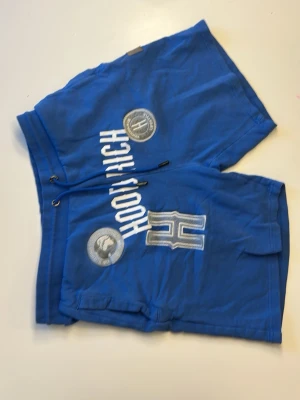 Blåa shorts med tryck och snörning - Snygga blåa shorts med vita tryck och stora bokstaven H framtill. Shortsen har snörning i midjan och flera coola detaljer som tryckta märken och fickor på sidorna. Perfekta för en avslappnad stil under varma dagar.