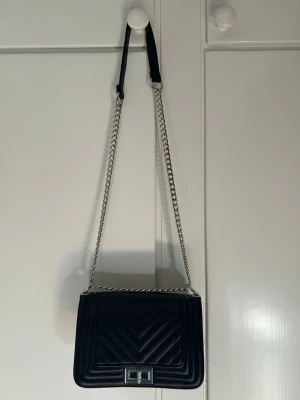 Svart quiltad axelväska med kedja - Snygg svart axelväska i quiltat mönster med silverfärgad kedjerem och metallspänne framtill. Väskan har en rektangulär form och justerbar axelrem med tygparti för extra komfort. Perfekt för dig som gillar stilrena accessoarer. Heelt oanvänd