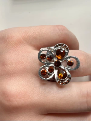Stor silver ring med oranga stenar formad som en blomma - Unik ring i silver med en stor blomformad front dekorerad med flera orange glittrande stenar. Ringen har en flexibel, stretchig ringdel som gör att den passar de flesta storlekar. Perfekt statement-accessoar för dig som gillar att sticka ut.