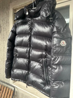 Svart dunjacka Moncler Maya - Moncler Maya storlek 2. Säljer en svart, glansig dunjacka från Moncler med huva och dragkedja framtill. Jackan har två snedställda fickor med dragkedja och Moncler-logga på ärmen.  Köpt på vinted helt ny och efter det är den sparsamt använd.  Qr kod samt NFC märke går allt att skanna.  Finns lite slitage på dragkedjan men annars i gott skick.