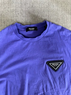 Lila t-shirt från Prada - Snygg lila t-shirt från Prada med klassisk rund halsringning och korta ärmar. På bröstet sitter en svart och vit Prada-logga i form av en triangel. Tillverkad i mjuk bomull som känns skön mot huden. Perfekt för dig som vill ha en stilren och trendig look.