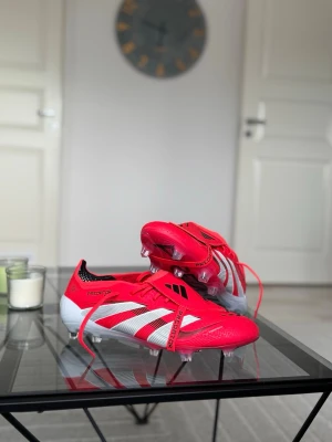 Adidas Predator röda fotbollsskor elit - Snygga Adidas Predator fotbollsskor i rött med vita och svarta detaljer. Skorna har klassisk snörning, mönstrad ovandel och tydliga Adidas-ränder på sidan. Yttersulan är vit med texten 'PREDATOR' och har fasta dobbar för spel på gräs.