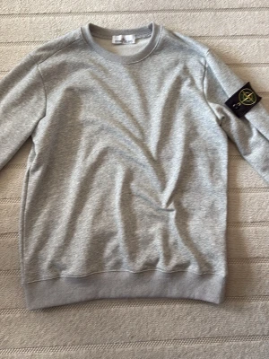 Grå sweatshirt från Stone Island - Snygg grå sweatshirt från Stone Island med klassisk rund hals och ribbade muddar. Tröjan har den ikoniska svarta patchen med gul och grön kompass på vänster ärm. Perfekt för en avslappnad och trendig stil.