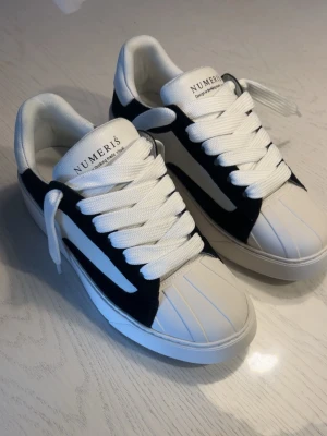Vita och svarta sneakers från Numeris - Snygga sneakers från Numeris i vitt med svarta detaljer och breda vita skosnören. Ovandelen har en unik paneldesign och sulan är vit och chunky. Skorna har en låg siluett och en modern streetstyle-vibe. Perfekta för dig som gillar stilrena men ändå iögonfallande sneakers.