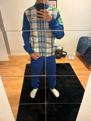 Blå och grön träningsdress - Säljer en retro träningsdress med blå byxor och jacka med rutigt mönster i blått, grönt, vitt och rött. Jackan har dragkedja och elastisk midja samt gröna detaljer på axlarna. Perfekt för sport eller chill. Materialet känns som polyester.