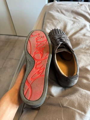 Gråa Christian Louboutin sneakers - Säljer ett par gråa sneakers från Christian Louboutin med klassisk röd sula och coola nitar över tån. Skorna är låga och har snörning samt är tillverkade i mocka för en lyxig känsla. Perfekta för dig som vill sticka ut med din stil. Storlek 42 passar 43 Ochså då min vän testa dom o sa dom satt bra 
