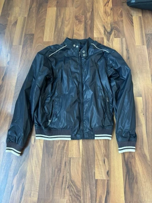 Marinblå vindjacka från Fred Perry - Snygg svart vindjacka från Fred Perry med vita detaljer på krage, muddar och axlar. Jackan har dragkedja framtill, två sidofickor med dragkedja och meshfoder på insidan. Perfekt för dig som gillar sportig och stilren look.