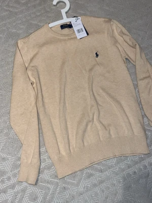 Beige stickad tröja från Polo Ralph Lauren - Klassisk beige stickad tröja från Polo Ralph Lauren med rund halsringning och diskret broderad logga på bröstet. Tröjan har långa ärmar och ribbade muddar vid ärmslut och nederkant. Perfekt basplagg för en stilren look.