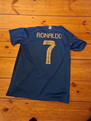 Blå Al Nassr Ronaldo matchtröja - Snygg blå matchtröja från Al Nassr med Ronaldo och nummer 7 på ryggen. Tröjan har guldfärgade detaljer, klubbmärke och sponsorloggor framtill. Tillverkad i ett lätt och ventilerande material, perfekt för fotbollsträning eller match.