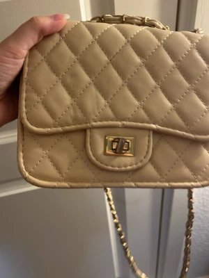 Beige quiltad axelväska med kedja - Snygg beige axelväska i quiltat mönster med guldfärgad kedjerem och vridlås. Väskan har en klassisk rektangulär form och är tillverkad i konstläder. Perfekt för dig som gillar stilrena accessoarer med en lyxig touch.