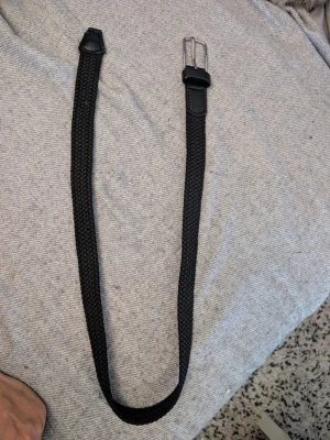 Svart flätat bälte från Kappahl - Snyggt svart flätat bälte från Kappahl i storlek 75. Bältet har en klassisk metallspänne och detaljer i skinnimitation. Perfekt för att ge din outfit en clean och stilren touch.