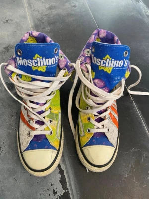  Moschino high-top sneakers - Unika high-top sneakers från Jeremy Scott med färgglad pop art-design. Skorna har inslag av blått, lila, grönt, orange och vitt med tryck som påminner om läskburkar. Tjock vit sula, vita snören och canvasmaterial. Perfekta för dig som vill sticka ut. Storlek 42 passar 41! Priset kan diskuteras