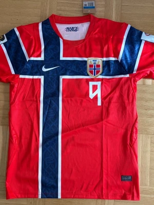 Norge fotbollströja Nike röd - Säljer en röd Norge fotbollströja från Nike med stort kors i blått och vitt, landslagets emblem på bröstet och nummer 9. På ryggen står det 'BRAUT HAALAND' och siffran 9. Perfekt för dig som älskar fotboll och vill stötta Norge.