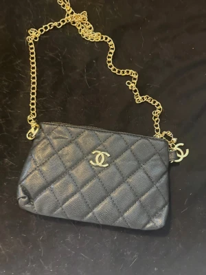 Svart axelväska/clutch med kedja - Snygg svart axelväska eller clutch om man vill  i läder med guldfärgad kedja och dragkedja. elegant design som ger en lyxig känsla. Perfekt för att bära det viktigaste med stil.