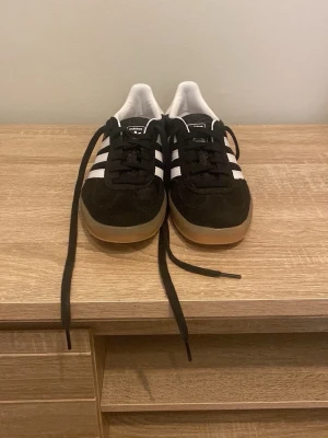 Adidas Gazelle svarta sneakers - Klassiska Adidas Gazelle sneakers i svart mocka med vita detaljer och gummisula. Skorna har de ikoniska tre vita ränderna på sidan, vit hälkappa och svart snörning. Perfekta för en clean och stilren look.