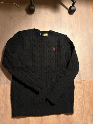 Svart kabelstickad tröja Polo Ralph Lauren - Kabelstickad svart tröja från Polo Ralph Lauren med rund halsringning och den klassiska röda logotypen broderad på bröstet. Tröjan har långa ärmar och ribbade muddar vid ärmslut och nederkant. Perfekt för lager på lager och stilren look.
