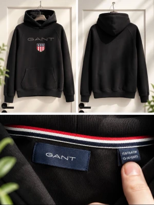 Gant Hoodie - Svart hoodie från GANT med stor ficka framtill, broderad GANT-text och sköldlogga på bröstet. Klassisk huva och ribbade muddar. Snygg detalj med röd, vit och marinblå rand på insidan av kragen. Perfekt för en chill och stilren look.
