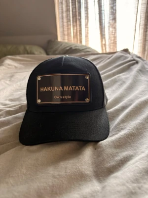 Svart keps med Hakuna Matata-plakett - Snygg svart keps med metallplakett framtill där det står 'HAKUNA MATATA Own style'. Kepsen har böjd skärm och mesh på sidorna och bak för extra ventilation. Perfekt för dig som vill ha en unik och chill stil.