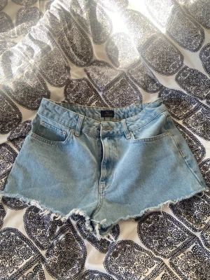Ljusblå jeansshorts - Säljer ett par ljusblå jeansshorts med fransig, rå kant och klassisk femficksdesign. Shortsen har bälteshällor och dragkedja framtill. Perfekta för varma dagar och en avslappnad stil. Från Nelly🦋🩵