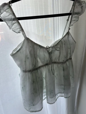 Ljusgrön blommig blus med volang HOLLISTER - Söt och luftig blus i ljusgrönt transparent tyg med små blommor, volangdetaljer på axelbanden och knyt framtill. Lite öppen i ryggen med en så fin knytning.  Perfekt till sommaren och ger en romantisk vibe till din outfit. 