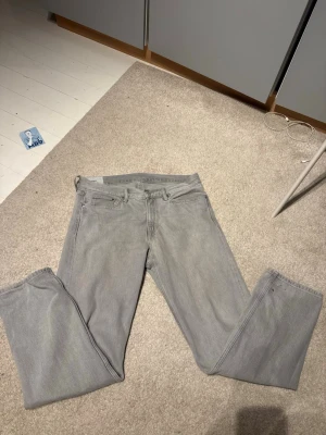 Ljusgrå raka jeans - Säljer ett par ljusgrå jeans med rak passform och klassisk femficksdesign. Jeansen har bälteshällor och stängs med dragkedja och knapp. Perfekta till en avslappnad stil och passar till det mesta. 33/32