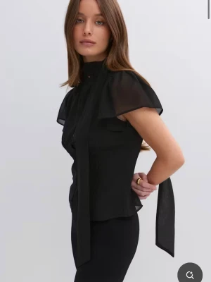 Svart blus med knytband i halsen - Stilren svart blus med korta, transparenta ärmar och högt knytband i halsen. Blusen har figurnära passform och lätt glansigt material som ger en elegant känsla. Perfekt för dig som gillar trendiga och snygga detaljer. Använd en gång. Helt slutsåld 