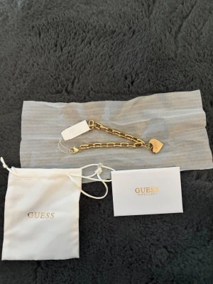 Guldfärgat armband med hjärthänge Guess - Snyggt guldfärgat armband från Guess med grova länkar och ett hjärtformat hänge där Guess-loggan är ingraverad. 