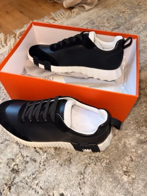 Svarta H sneakers med vit sula - Snygga svarta sneakers med chunky vit sula och svarta detaljer. Skorna har låga skaft, svarta skosnören och en unik såldesign med räfflad kant. Tillverkade i läder och mocka för en exklusiv känsla. Perfekta för dig som vill sticka ut.