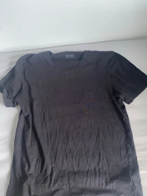 Svart t-shirt från BOSS - Snygg svart t-shirt från Hugo boss med diskret logga broderad på bröstet. Klassisk rund hals och korta ärmar. Tillverkad i mjukt bomullsmaterial som känns skönt mot huden. Perfekt basplagg för en clean och stilren look.