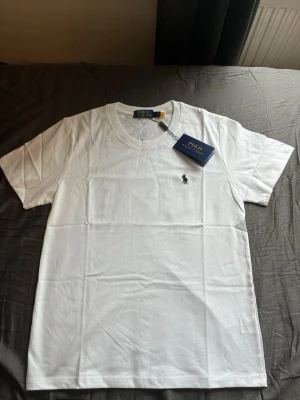 Ralph Lauren T-shirt  - Klassisk vit t-shirt från Polo Ralph Lauren med rund halsringning och den ikoniska broderade loggan på bröstet. Tillverkad i mjuk bomull för en skön och stilren look. Perfekt basplagg för alla tillfällen.