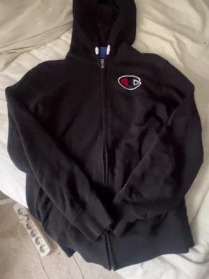 Svart zip-hoodie från Champion - Säljer en svart zip-hoodie från Champion med stor huva och det klassiska Champion-logotypen broderad på bröstet. Hoodien har dragkedja framtill och ribbade muddar vid ärmar och nederkant. Storlek är 11/12 years men kan sitta som en xxs/xs i dam modell
