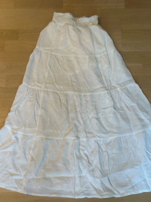 Vit lång kjol med smock i midjan - Luftig vit långkjol med smockad midja och flera volanger. Kjolen är gjord i ett tunt, mjukt bomullstyg som passar perfekt till varma dagar. Enkel och stilren design som är lätt att matcha med olika toppar.
