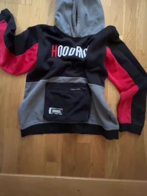Svart, grå och röd hoodie med tryck - Säljer en hoodie i svart, grå och röd färg med stor ficka framtill och texten 'HOODRICH' tryckt på bröstet. Huvan är grå och ärmarna har röda detaljer. Perfekt för dig som gillar streetwear och vill sticka ut lite extra.