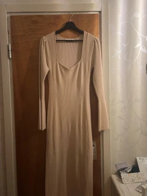 Beige ribbad långklänning - Säljer en beige ribbad långklänning med långärmad design och v-ringad hals. Klänningen har en kroppsnära passform och är tillverkad i ett stretchigt material som ger en snygg siluett. Perfekt för dig som gillar stilrena och trendiga plagg.