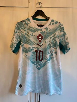 Portugal fotbollströja Puma nr 10 - Snygg Portugal fotbollströja från Puma med nummer 10 och namnet R. Leão på ryggen. Tröjan är vit med turkosa och ljusblå mönster, har korta ärmar och rund hals. Perfekt för dig som vill stötta landslaget eller spela själv.