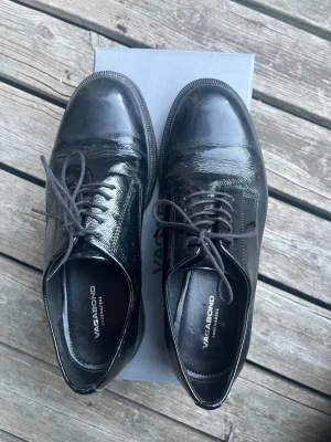 Svarta lackloafers från Vagabond - Säljer ett par svarta loafers från Vagabond i glansigt lackmaterial. Skorna har klassisk rund tå, snörning framtill och låg klack. Skorna är lätt använda (ca 10ggr) men tyvärr har jag insett att mina fötter är lite för breda för dem, älskar skorna annars. Skokartong medföljer! Originalpris 1400 kr, köptes på rea för 850 kr