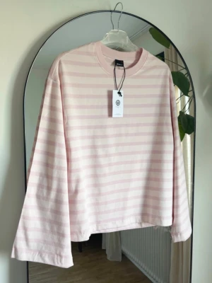 Ljusrosa och vit randig långärmad topp - En söt långärmad topp med ljusrosa och vita ränder. Toppen har en avslappnad passform och är tillverkad i mjuk bomull. Perfekt för dig som gillar en enkel men stilren look.