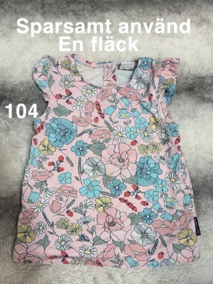 Blommig topp med volangärm - Söt topp i ljusrosa med färgglatt blommönster i blått, gult och vitt. Toppen har volangdetaljer vid ärmarna och är ärmlös. Perfekt för vår och sommar med lekfulla design.🌸Sparsamt använd. En fläck. Storlek 104 