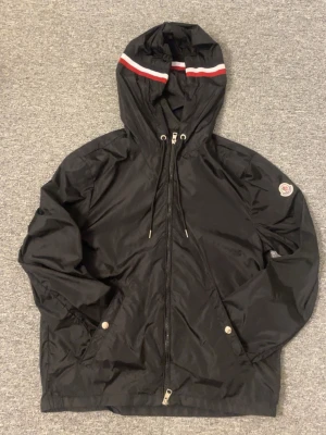 Moncler windbreaker - Snygg svart vindjacka med huva och dragsko. Jackan har dragkedja framtill, två fickor med tryckknappar och en vit/röd rand på huvan. Materialet är glansigt och lätt, perfekt för blåsiga dagar. Liten patch på ärmen för extra detalj.