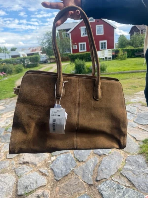 Brun mocka toteväska  - Snygg väska i brun mocka från NAKD. Väskan har en stilren design med dragkedja upptill och praktiska sidofack. Helt ny med lapp kvar, sista bilden visar från NAKD hemsida. Det har kommit lite vattenstänk så de finns lite fläckar på men går att få bort med tex en mockaborste. Första bilden är den lite orättvis i färgen 